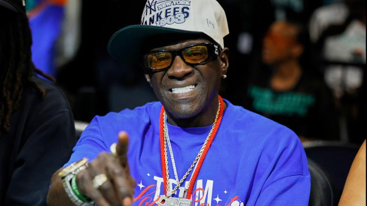 Flavor Flav holding a clock, smiling, with Atlanta Dream fans cheering in the background.
